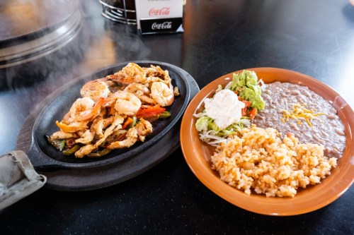 Sizzling shrimp fajitas on a hot plate, served with rice, refried beans, guacamole, and salad on a separate plate.