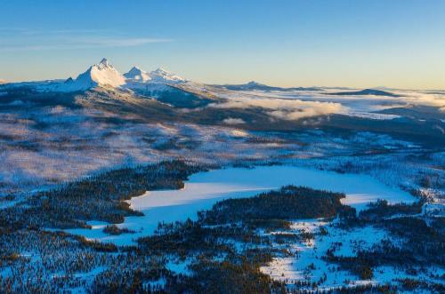 Snow-covered mountains rise above a frozen lake, surrounded by a forest under a clear blue sky.