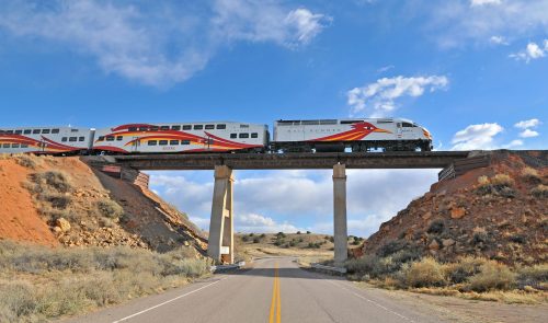A colorful train crosses a concrete bridge over a road, surrounded by rocky terrain and blue skies.