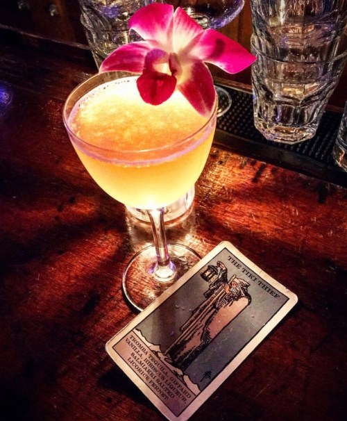 A cocktail garnished with a pink flower sits on a bar next to a tarot card titled "The Hanged Man."