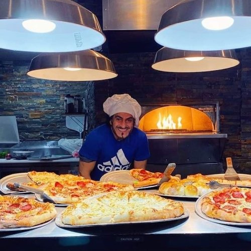 A chef in a hat smiles behind a display of large pizzas, with a wood-fired oven glowing in the background.