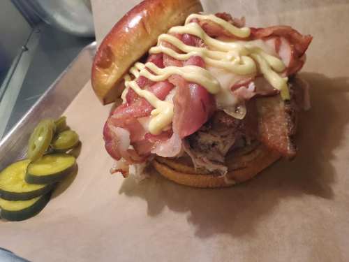 A hearty sandwich piled with bacon, pulled pork, and drizzled with mayonnaise, served with pickles on the side.