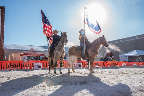 Two riders on horseback hold American and Texas flags, with a bright sun shining in the background during an outdoor event.