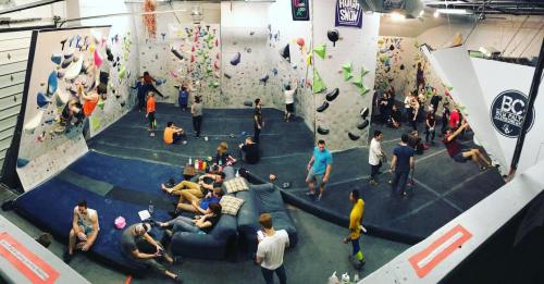 A busy indoor bouldering gym with climbers on walls and people relaxing on couches in a social area.