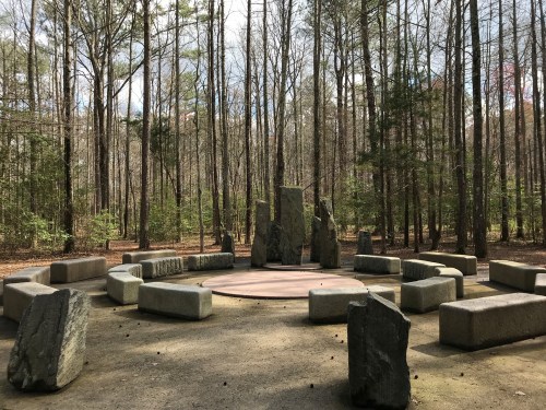 A circular stone arrangement in a wooded area, surrounded by tall trees and a clear sky.