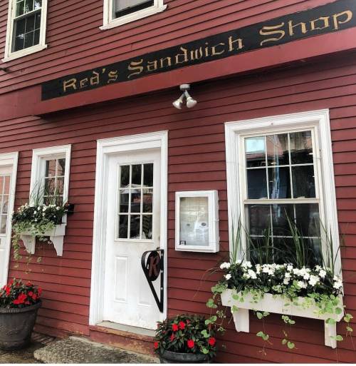 A red building with a sign reading "Red's Sandwich Shop," featuring flower boxes and a white door.