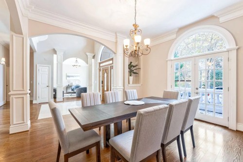Elegant dining room with a large table, upholstered chairs, and arched windows, leading to a bright living area.