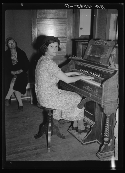 A woman in a floral dress plays an organ while another woman sits in the background, both in a vintage interior setting.