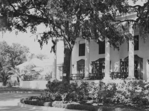 A historic mansion with columns, surrounded by trees and lush greenery, in black and white.