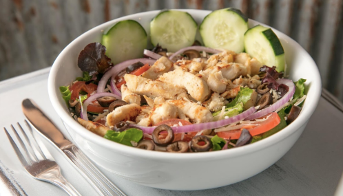 A fresh salad with grilled chicken, cucumbers, olives, tomatoes, and red onions in a white bowl. Fork and knife beside it.