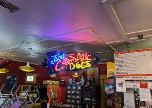 Neon sign for "Jack's Cosmic Dogs" inside a colorful diner with arcade games and a menu board.