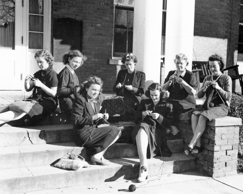A group of seven women sitting on steps, knitting together, smiling, in a vintage black-and-white photo.