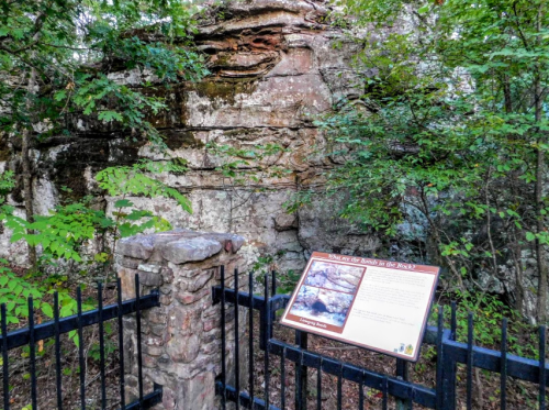 A stone pillar and informational sign near a rocky cliff, surrounded by trees in a natural setting.