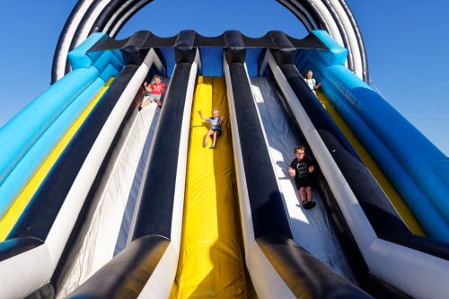 Four children sliding down a large inflatable slide on a sunny day, with blue sky in the background.