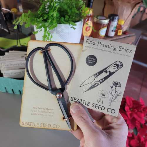 A hand holding fine pruning snips and a packaging card from Seattle Seed Co, with plants in the background.