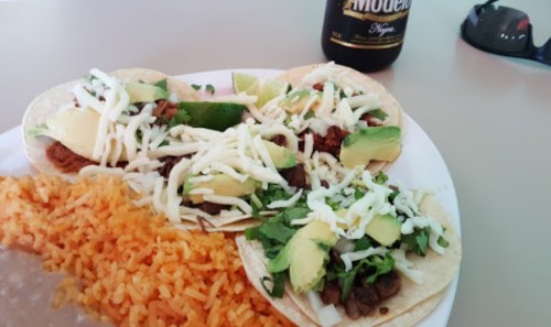 Three tacos topped with cheese, avocado, and lime, served with a side of orange rice and a bottle of beer.