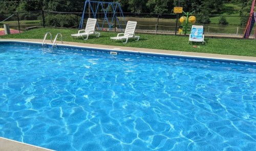 A clear blue swimming pool with lounge chairs nearby, surrounded by grass and a playground in the background.