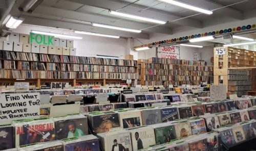A record store interior filled with vinyl records, organized by genre, with signs for different sections and prices.