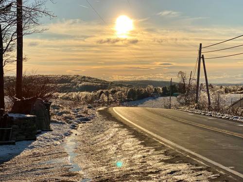A winter landscape at sunset, featuring a snowy road, trees, and a glowing sun above distant hills.