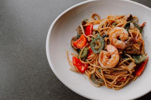 A bowl of shrimp pasta with vegetables, including bell peppers and mushrooms, on a dark countertop.
