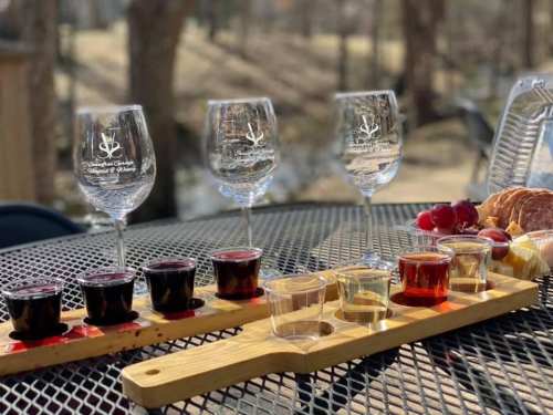 A wooden flight board with various colorful drink samples, accompanied by wine glasses and snacks on a table outdoors.