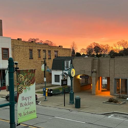 A sunset casts a warm glow over a small town street, with a holiday sign and buildings in view.