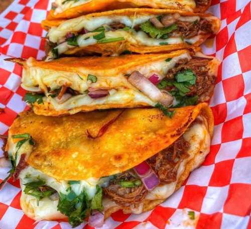 A close-up of crispy tacos filled with meat, cheese, onions, and cilantro, served in a red and white checkered basket.