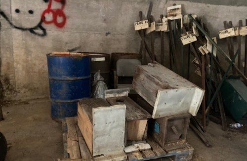 A cluttered storage area with wooden crates, barrels, and metal containers against a concrete wall.