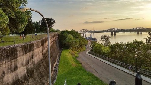 A scenic view of a riverbank with a path, trees, and a bridge in the background during sunset.