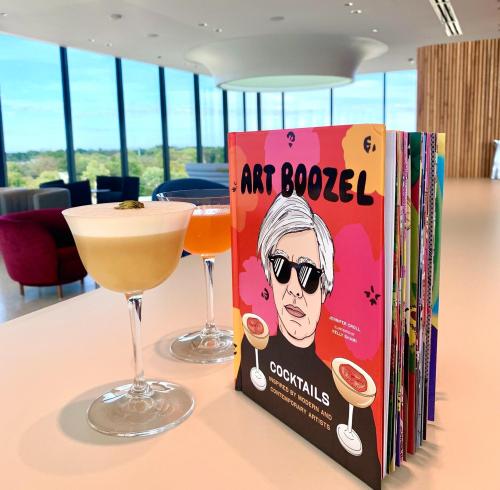 A cocktail book titled "Art Boozel" with two colorful cocktails on a table, overlooking a scenic view through large windows.