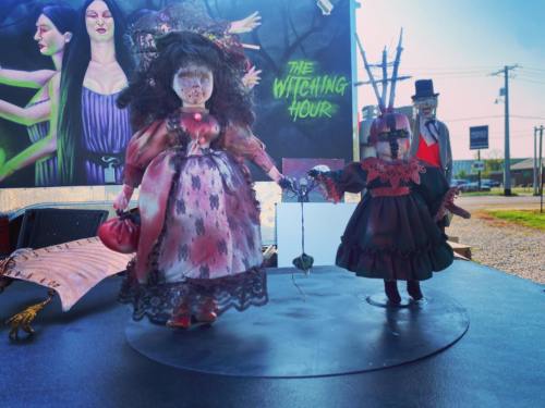 Two vintage dolls in elaborate costumes, holding hands, displayed against a colorful Halloween-themed backdrop.