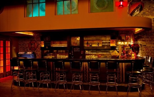 A dimly lit bar with a sleek counter, high stools, and colorful lighting, featuring vintage decor and a jukebox.