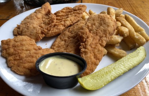 Plate of crispy fried chicken tenders, golden fries, a pickle slice, and a small cup of dipping sauce.
