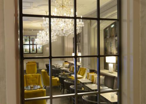 View through a window into a stylish lounge with yellow chairs, elegant chandeliers, and modern decor.