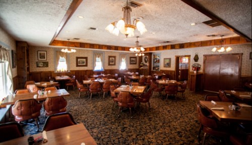 Cozy restaurant interior with wooden walls, patterned carpet, and empty tables set for dining under warm lighting.