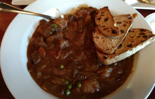 A bowl of hearty beef stew with vegetables, served with slices of toasted bread on the side.