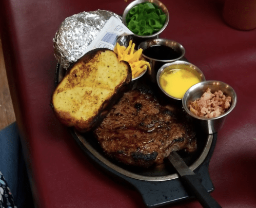 A sizzling steak on a platter with garlic bread, baked potato, cheese, green onions, and sauces.
