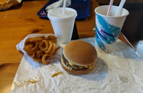 A burger with cheese, curly fries, and two drinks on a table with a paper wrapper.