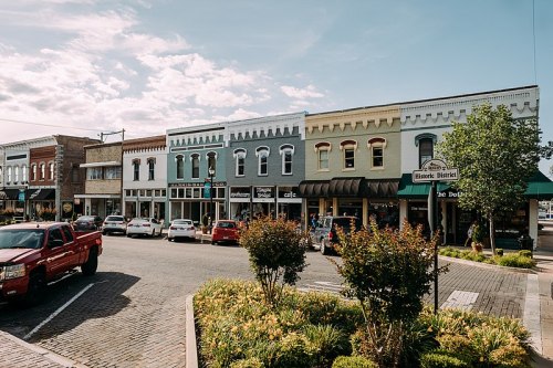 Charming small-town street with colorful storefronts, parked cars, and greenery under a blue sky.