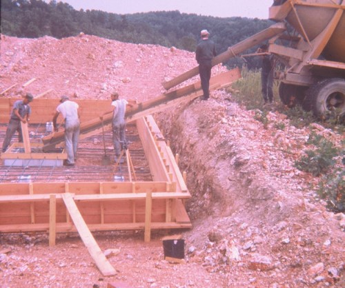 Workers pouring concrete into a foundation frame at a construction site, with machinery and materials in the background.