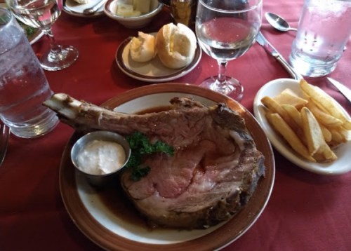 A plate with a large ribeye steak, a side of fries, dipping sauce, and bread rolls on a red tablecloth.