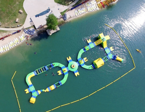 Aerial view of a colorful water obstacle course on a lake, surrounded by kayaks and people enjoying the water.