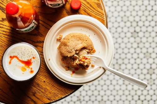 A partially eaten fried dish on a plate, accompanied by hot sauce and a drink, on a wooden table with a tiled surface.