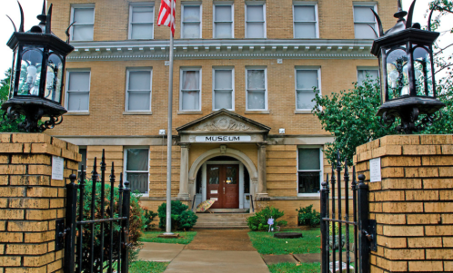 Historic museum building with an American flag, flanked by lanterns and surrounded by greenery.