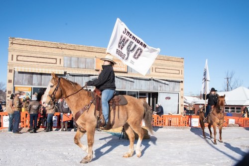 A rider on a horse carries a flag during a winter rodeo event in a small town, with spectators in the background.