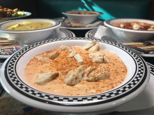 A bowl of creamy crab soup topped with crab meat and spices, surrounded by other dishes in the background.