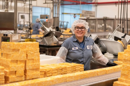 A smiling worker in a cheese factory stands by stacks of cheese, wearing a hairnet and gloves.