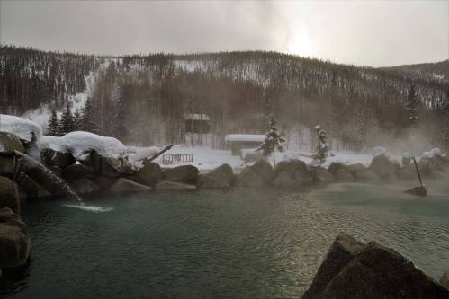 A serene hot spring surrounded by snowy mountains and mist, with rocks and trees in the foreground.