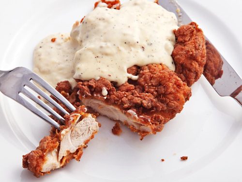 A plate of fried chicken topped with creamy gravy, with a fork and knife ready to serve.