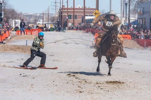 A cowboy on a horse races past a snowboarder being pulled on a snowy street, with a crowd watching in the background.